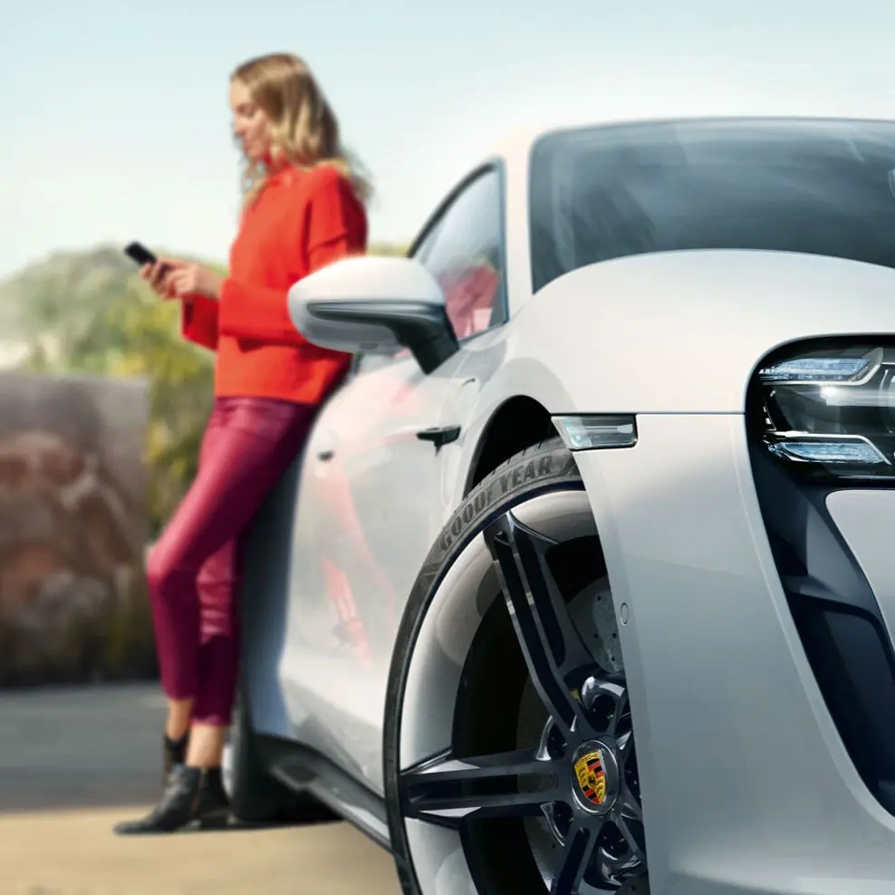 Abbild einer Frau die an einem Porsche lehnt und mit ihrem Smartphone die digitalen Porsche Services nutzt.